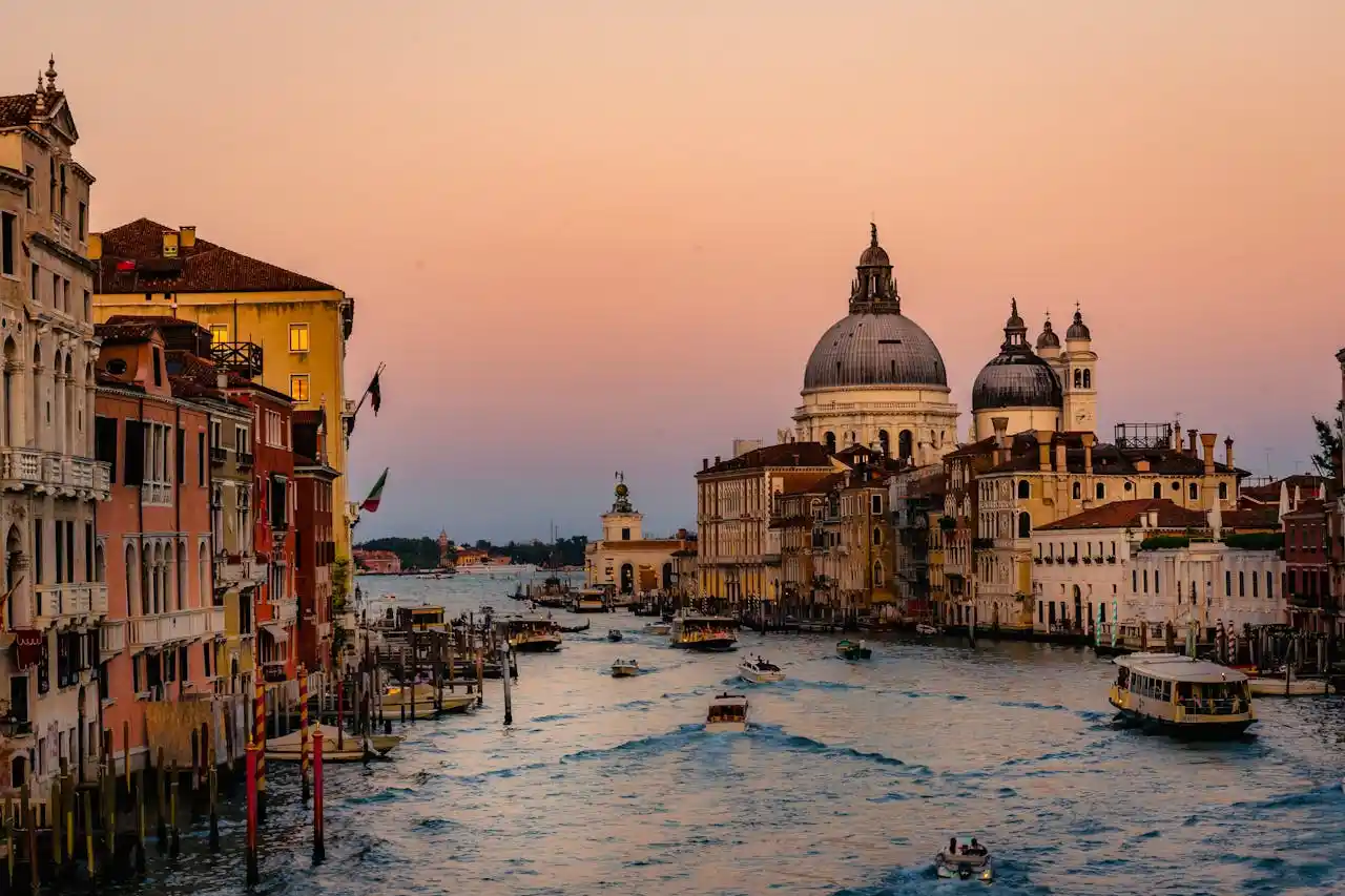 A picturesque view of Venice's Grand Canal at sunset, featuring boats traversing the water, flanked by historical buildings with colorful facades. The domed Santa Maria della Salute church stands prominently against a pastel pink and orange sky.