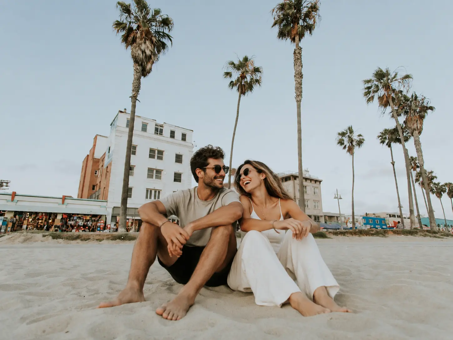 A man and woman sit barefoot on a sandy beach, smiling at each other. Palm trees and buildings are in the background under a clear sky.