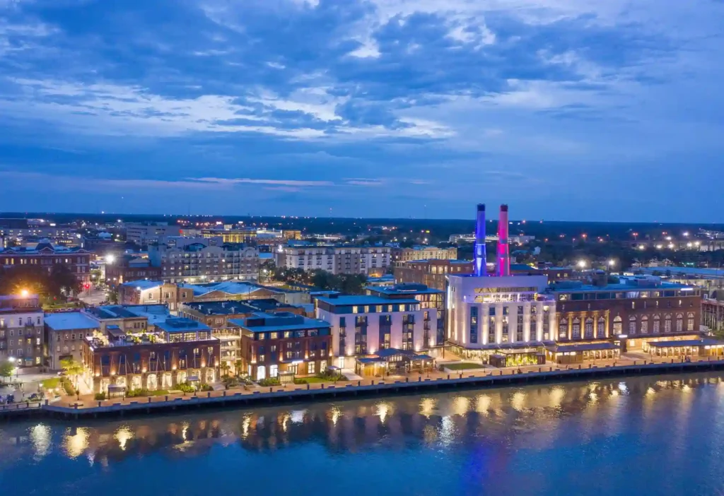 Aerial view of a waterfront cityscape at dusk. Buildings are illuminated, with a prominent structure featuring lit smokestacks in purple and pink. Reflections shimmer on the water, and a partly cloudy sky adds to the serene atmosphere.