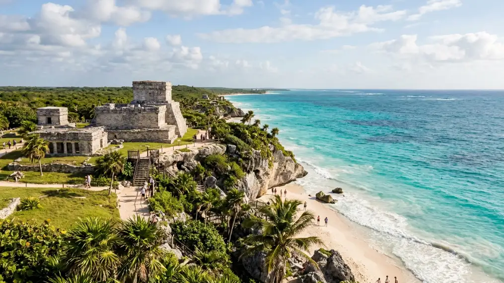 Ancient Mayan ruins sit atop a cliff overlooking a white sandy beach and turquoise ocean, surrounded by lush greenery and palm trees under a partly cloudy sky. Tourists walk along the shoreline below. and this is one of the Best things to do in Tulum for tourists.