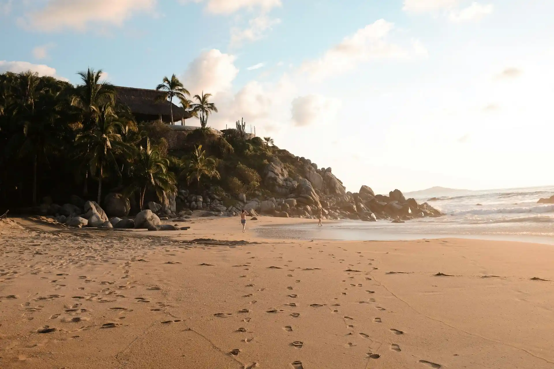A sandy beach with footprints leads to a rocky hill covered in palm trees and a house. Two people walk near the waters edge under a partly cloudy sky during sunset. Waves gently hit the shore.